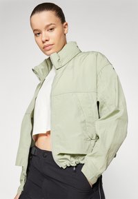 Vaaleanvihreä bomber-takki, jossa on vetoketju edessä, nepparikaulus, joustava helma ja sivutaskut. Päällä valkoinen crop-top.