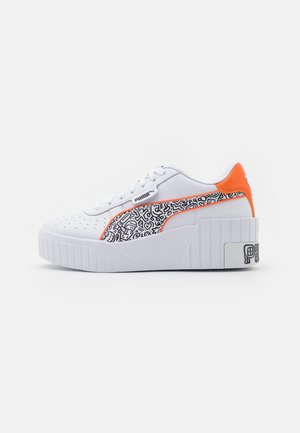 Baskets Puma blanches avec semelle épaisse, bande à motif abstrait noir et blanc, et accent orange au talon sur un fond clair uni.