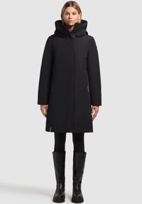 Manteau noir imperméable avec capuche, fermeture éclair, deux poches latérales et coupe droite. Porté sur des leggings noirs et des bottes montantes.