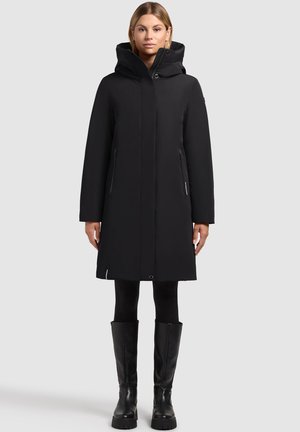 Zwarte waterdichte jas met een capuchon, ritssluiting, twee zijzakken en een rechte snit. Gedragen over zwarte leggings en kniehoge laarzen.