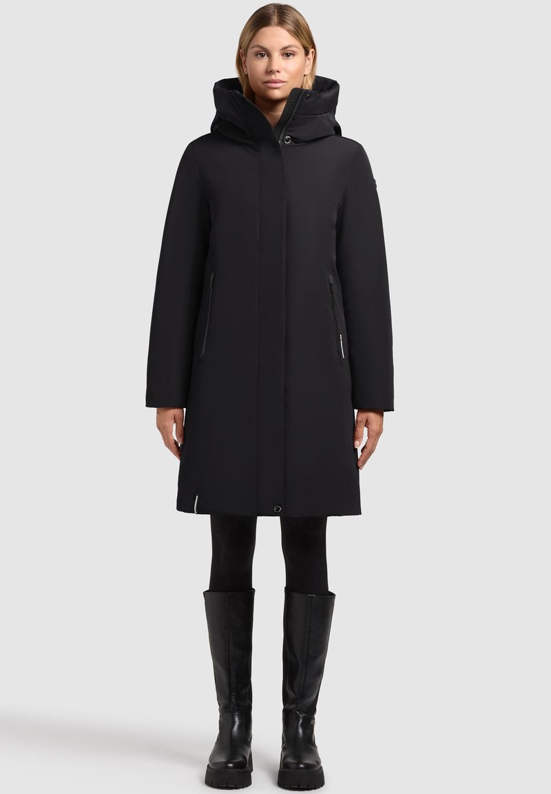 Manteau noir imperméable avec capuche, fermeture éclair, deux poches latérales et coupe droite. Porté sur des leggings noirs et des bottes montantes.