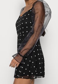 Czarna krótka sukienka w białe kropki. Posiada przezroczyste, bufiaste rękawy z elastycznymi mankietami. Zwiewny dół spódnicy i dekolt w kształcie litery V.