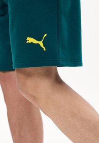 Κοντινό πλάνο στο πόδι ενός ατόμου που φοράει σκούρα πράσινα σορτς με μικρό κίτρινο λογότυπο Puma κοντά στη ραφή.