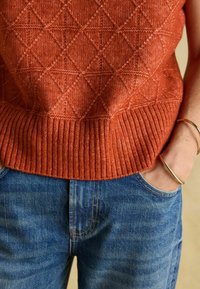 Inimene kannab roostevärvi kudumit, millel on teemandimuster, ja siniseid teksaseid, käsi taskus, randmel kuldne käevõru.