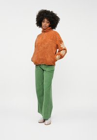 oranžový fleece pulóver s krátkym zipsom a bielou kvetinovou výšivkou na rukávu, skombinovaný so zelenými zamatovými širokými nohavicami a svetlými topánkami.