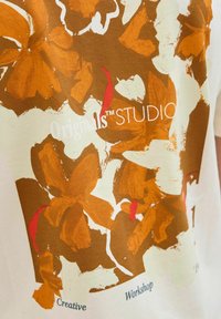 Trumpas rankovėmis baltas marškinėliai su rudais floriniais motyvais, raudonais ir kreminiais akcentais, taip pat užrašas "Originals™ STUDIO".