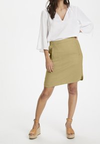Jupe portefeuille kaki avec poches latérales, confectionnée dans un tissu léger. Associée à un chemisier blanc ample et des sandales compensées tan.