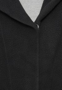 Casaco de fleece preto com uma superfície texturizada, apresentando um colarinho tipo lapela e um fecho de fecho éclair prateado na frente. Tecido macio e quente.