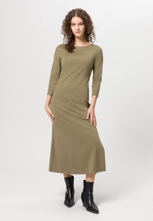 Robe longue à manches longues de couleur vert olive avec un col rond, texture lisse et une longueur moyenne, associée à des bottines noires.