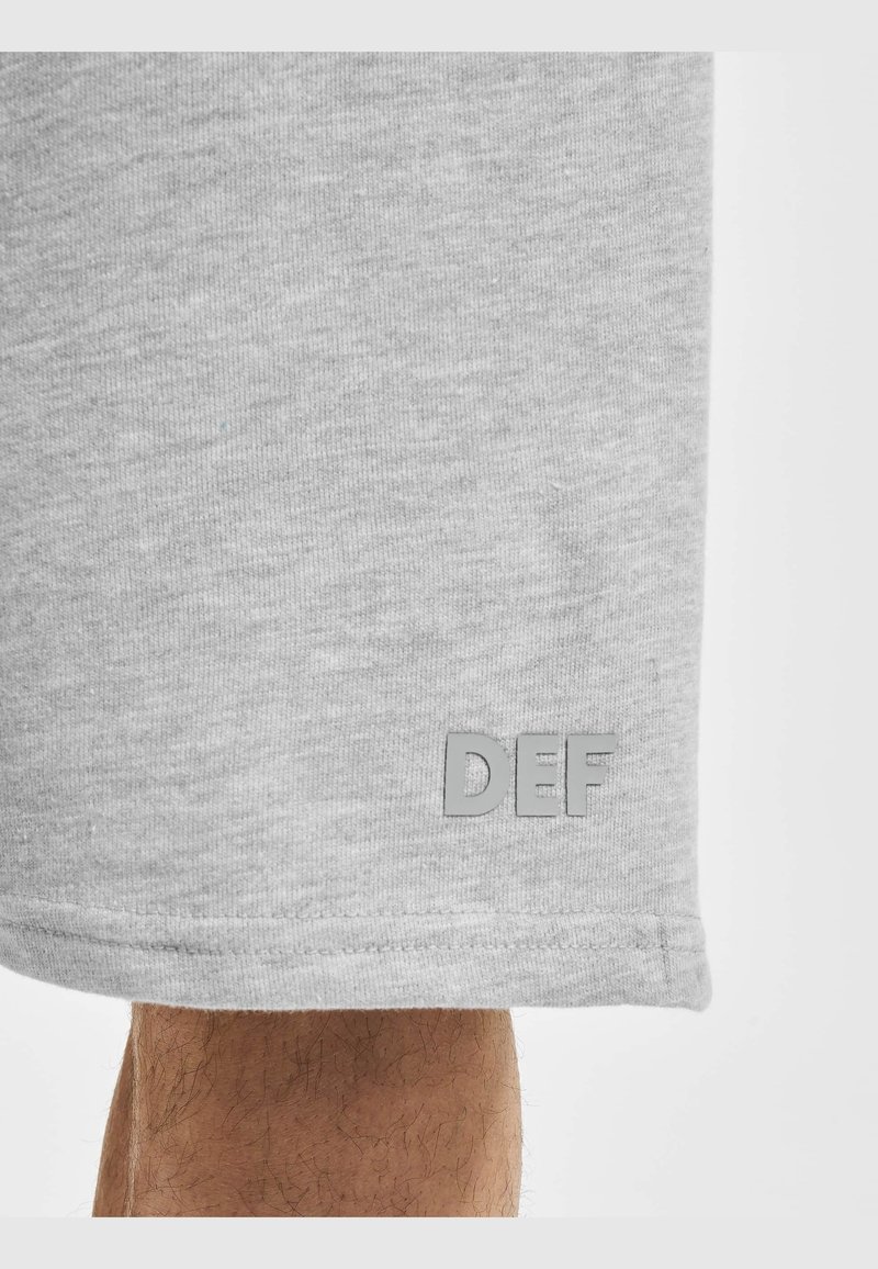 Shorts en tissu gris avec le logo "DEF" près de l'ourlet inférieur au-dessus d'une partie visible de la jambe d'une personne avec des poils.