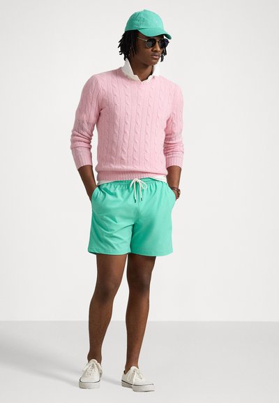 Suéter rosa em ponto de malha, calções de banho verde-menta, sapatos brancos e uma capa mint. Acessórios incluem óculos de sol pretos, exibindo um traje de verão.