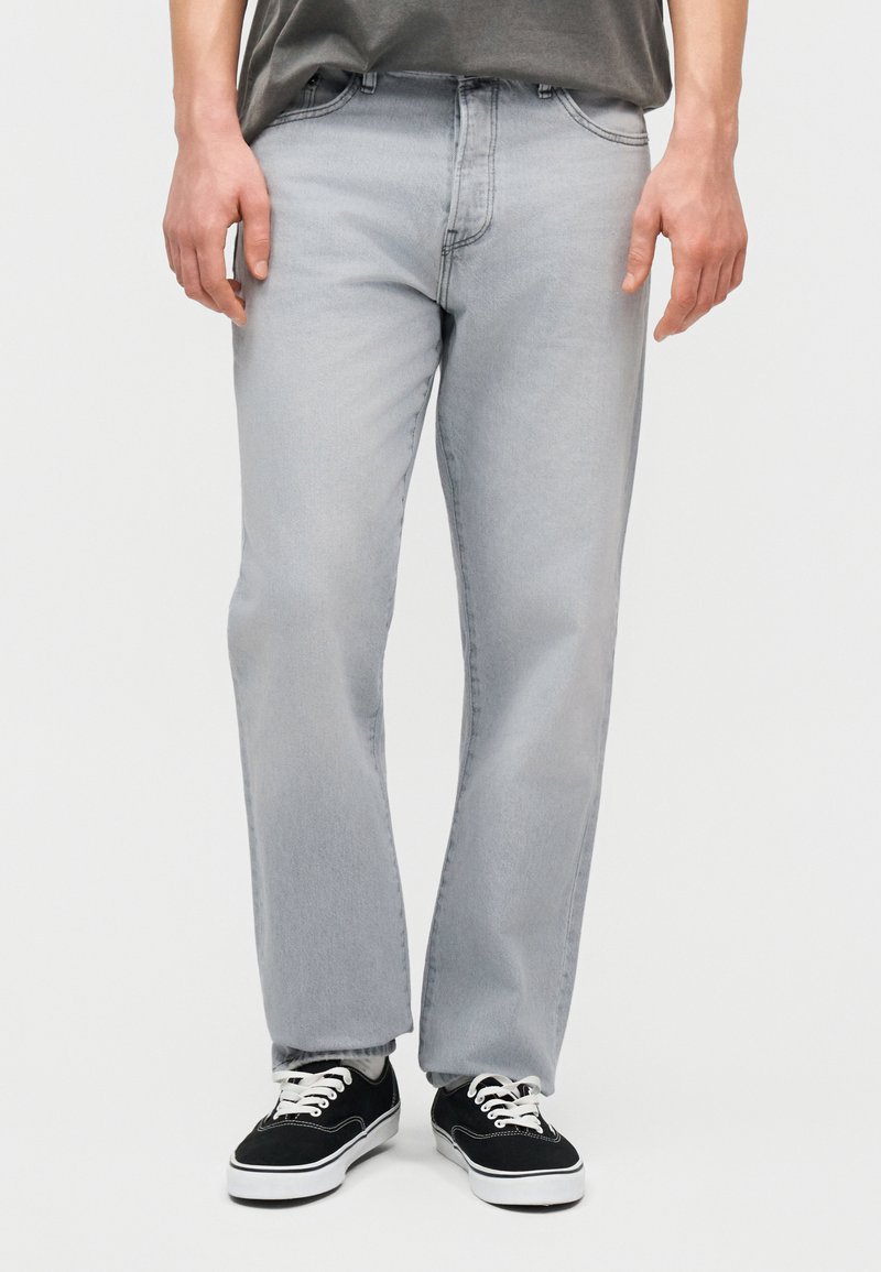 Άτομο που φοράει ανοιχτό γκρι τζιν με ίσια μπατζάκια και μαύρα χαμηλά παπούτσια με λευκά κορδόνια, στέκεται μπροστά σε ένα απλό φόντο.