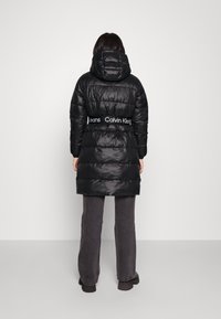 Svart puffjacka med huva, med quiltat mönster och elastisk midja med "Calvin Klein Jeans"-logotyp. Bärs över grå byxor.