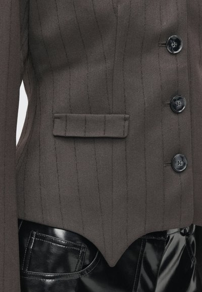 Sötétbarna, szabott blézer függőleges csíkokkal, egyetlen mellzsebbel, két fekete gombbal és ívelt aljjal, fényes fekete nadrággal párosítva.