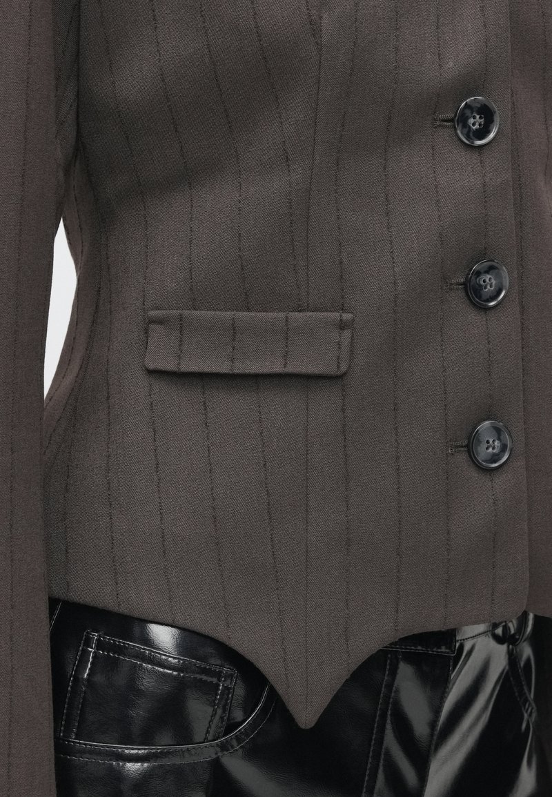 Mørkebrun skræddersyet blazer med vertikale striber, der har en enkelt brystlomme, to sorte knapper og en buet kant, parret med skinnende sorte bukser.