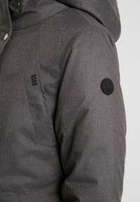 Giacca isolante grigia con motivo a spina di pesce, caratterizzata da una toppa nera con logo sul braccio e una zona spalle testurizzata.