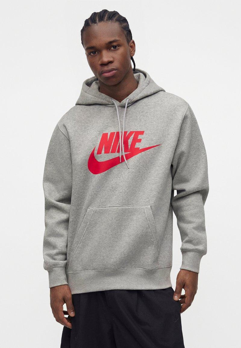 Hombre con el cabello trenzado que lleva una sudadera gris con un gran logo rojo de Nike y pantalones negros, de pie frente a un fondo liso.