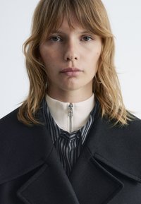 Black wool coat with large lapels over a striped shirt and cream mock neck. Features a front zipper and wrapped hair. Clear skin tone.