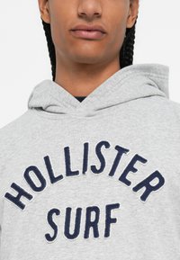 Osoba koja nosi svijetlosivi duksericu s tamnoplavi "HOLLISTER SURF" natpisom na prsima, tamna pletenica vidljiva.