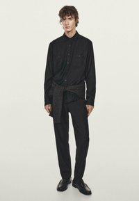 Schwarzes Hemd mit Knöpfen und zwei vorderen Taschen, kombiniert mit lockeren schwarzen Hosen. Ein dunkelgrauer Pullover, der um die Taille gebunden ist, und schwarze Schuhe.