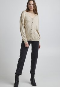 Cardigan beige a maglia con scollo a V, maniche lunghe e bottoni neri, abbinato a jeans neri con orlo frangiato e stivaletti.
