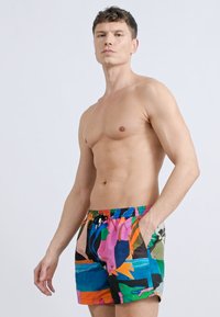 Άνδρας χωρίς πουκάμισο με κοντά μαλλιά, φοράει πολύχρωμα μαγιό με αφηρημένο σχέδιο, στέκεται με το ένα χέρι στην τσέπη μπροστά σε απλό φόντο.