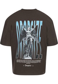 Brun oversized t-skjorte med et grafisk trykk av en engel statue med utstrakte vinger, tekst i blått over og under designet.