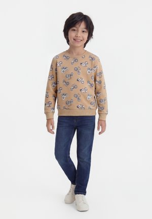 Enfant portant un sweat-shirt beige avec un motif de tracteur, un jean bleu et des baskets blanches, debout et souriant sur un fond uni.
