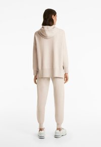 Personne portant un hoodie beige et un jogging assorti avec des baskets blanches, standing de dos devant un fond blanc uni.