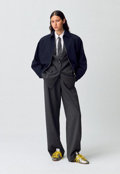 Chaqueta bomber con cremallera en navy, traje gris a rayas, camisa blanca, corbata en blanco y negro, zapatillas plateadas con cordones amarillos. Mezcla destacada de estilo casual y formal.