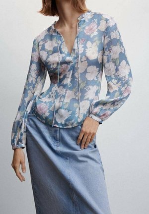 Vrouw draagt een blauwe bloes met bloemenprint, lange mouwen met een V-hals en een lichtblauwe spijkerrok tegen een effen achtergrond.