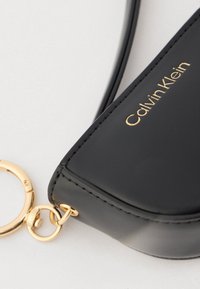 Μαύρο δερμάτινο πορτοφόλι κλειδιών Calvin Klein με χρυσό κρίκο και κούμπωμα σε λευκό φόντο.