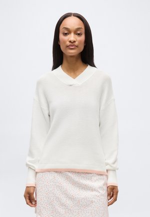 Femme aux cheveux longs et raides portant un pull en tricot blanc avec des bordures roses et une jupe blanche à petits motifs de feuilles roses, se tenant devant un fond uni.