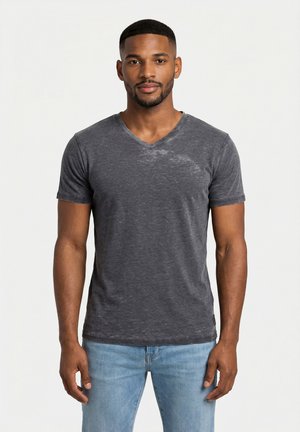 Uomo con capelli corti e barba che indossa una T-shirt scollo a V aderente grigio scuro e jeans azzurri, in piedi davanti a uno sfondo semplice.