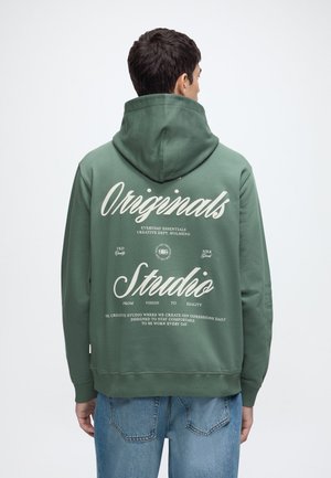 Hombre con sudadera verde con texto blanco "Originals Studio" y frases más pequeñas en la espalda, combinado con vaqueros azules, de espaldas a la cámara.
