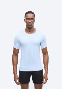 Mand iført en lyseblå, tætsiddende kortærmet skjorte og sorte shorts, stående med fronten mod kameraet og et neutralt udtryk.