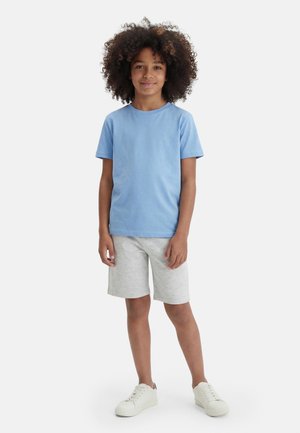 Kind mit lockigem Haar steht, trägt ein hellblaues Kurzarmhemd, graue Shorts und weiße Turnschuhe vor einem einfarbigen Hintergrund.