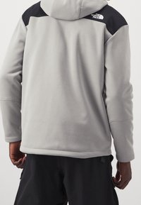 Graue Fleecejacke mit einem schwarzen Schulterbereich, mit Kapuze und sichtbaren Nähten, getragen über dunklen Hosen.