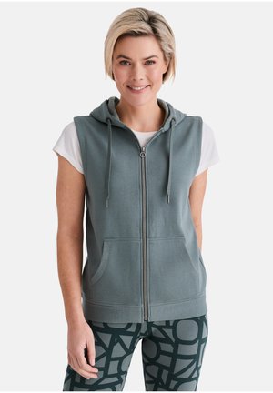 Lächelnde Frau mit kurzen blonden Haaren, die einen ärmellosen Kapuzenpulli mit Reißverschluss über einem weißen T-Shirt und gemusterten Leggings trägt, steht vor einem schlichten Hintergrund.