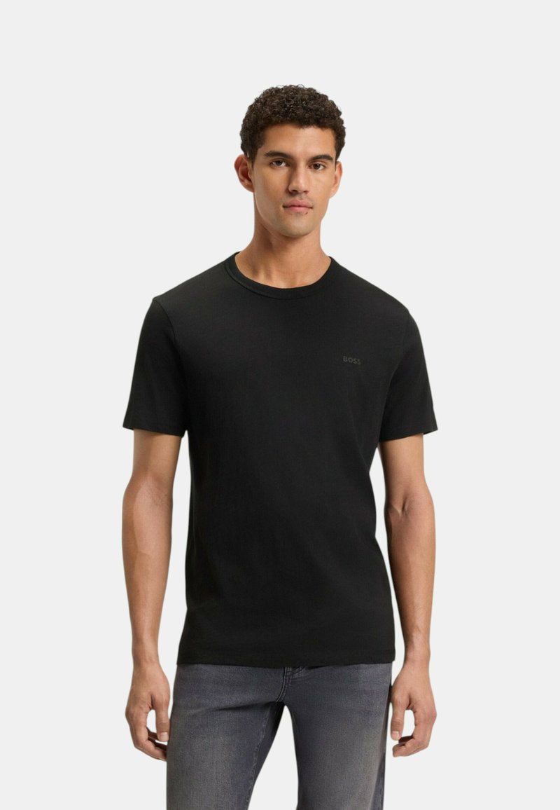Homme aux cheveux courts et bouclés portant un t-shirt noir uni à manches courtes et un jean gris, debout devant un fond blanc.