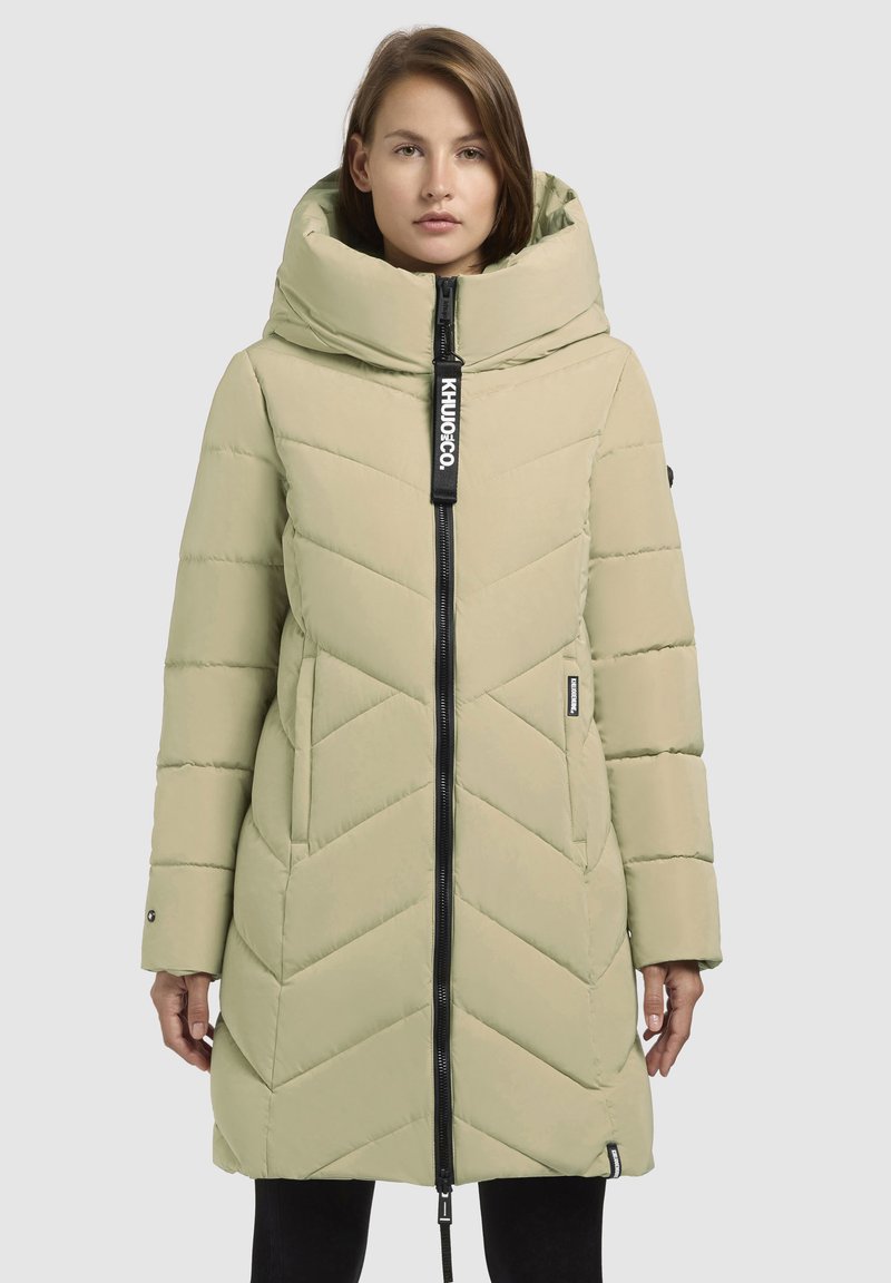 Abrigo acolchado verde claro con cuello alto, costura en forma de chevron, cremallera completa, etiqueta de marca y bolsillos laterales. Textura suave.
