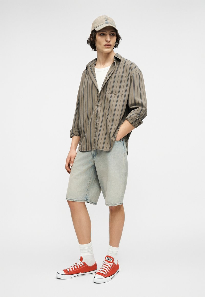 Junger Mann, der ein beige gestreiftes Hemd, helle Jeansshorts, eine beige Kappe, weiße Socken und rote Sneaker mit niedrigem Schaft trägt, steht mit einer Hand in der Hosentasche.