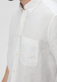 Man in een wit linnen korte mouwen blouse met een borstzak en een button-down kraag, gezien vanaf de zijkant.