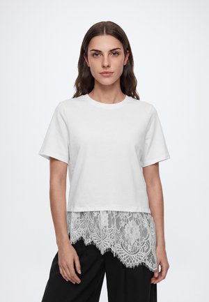 Femme aux longs cheveux bruns portant un haut blanc à manches courtes avec un détail en dentelle florale transparente à l'ourlet et un pantalon noir, faisant face à l'avant.