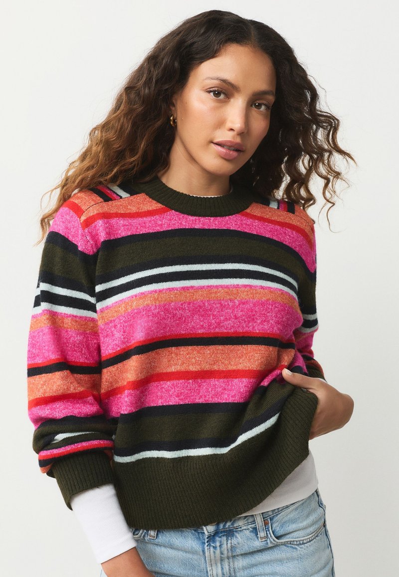 Kvinde med bølget hår iført en flerfarvet stribet sweater i orange, pink, grøn og sort, kombineret med lyseblå jeans, mod en ensfarvet baggrund.