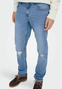 Vaqueros de mezclilla azul claro con corte slim, que presentan desgarros deshilachados en las rodillas y un ligero desgaste, combinados con zapatos marrones de cordones.