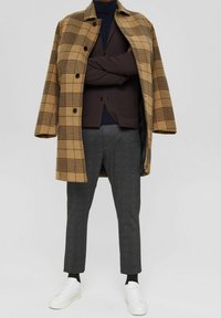 Brauner und gelber karierten Übermantel, graue Hose, marineblaues Hemd, dunkelbraune Weste, weiße Sneakers. Strukturiertes Gewebe mit Knopfverschlüssen.