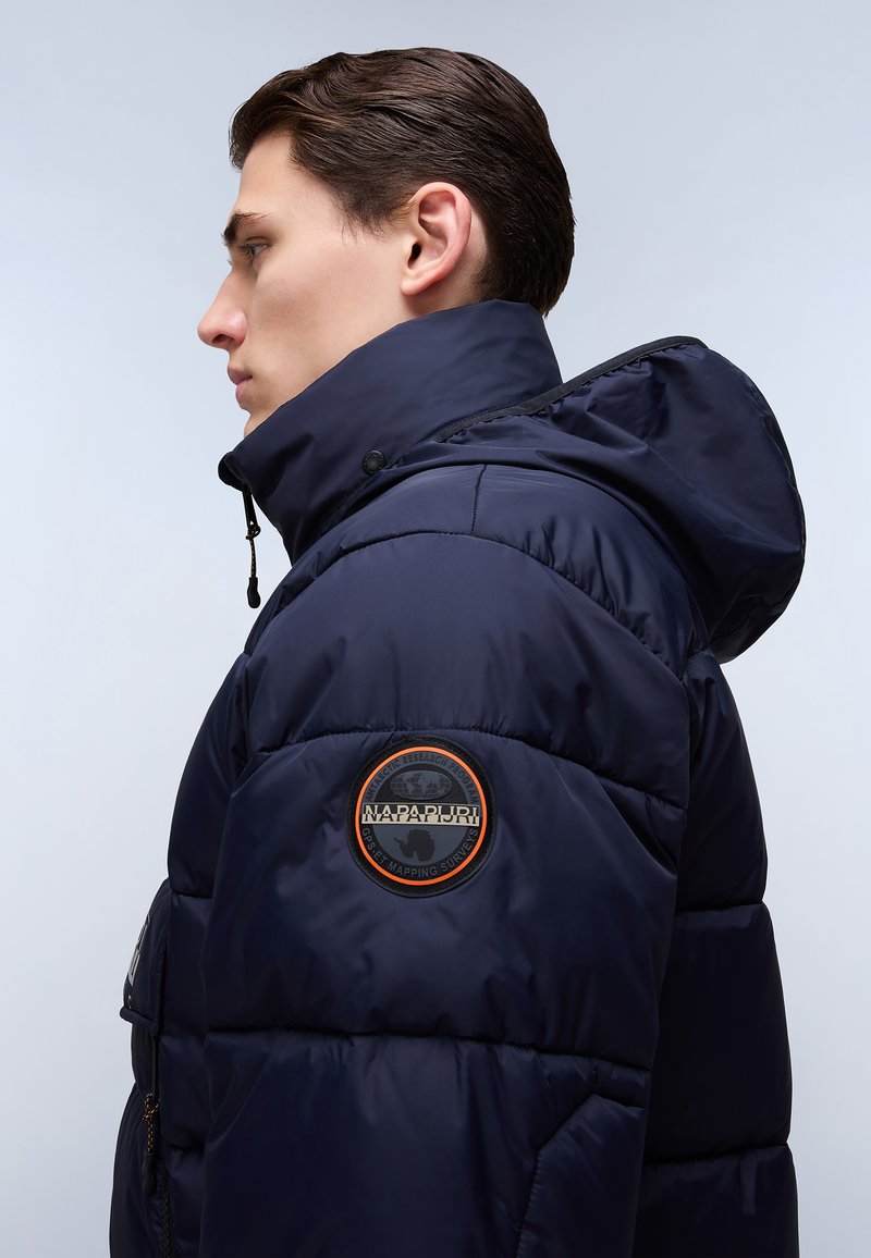 Marineblauwe pufferjas met een hoge kraag en capuchon. Bevat horizontale gestikte panels en een ronde logopatch op de linker mouw.