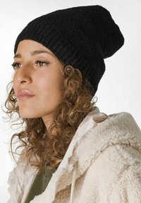 Svart stickad mössa med en avslappnad design, med ett texturerat mönster. Modellen bär en krämfärgad fluffig jacka under.