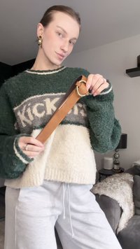 Oseba v zeleno-belem puhastem puloverju in belih hlačah drži rjav usnjen pas z okroglim zaponko v notranjosti.